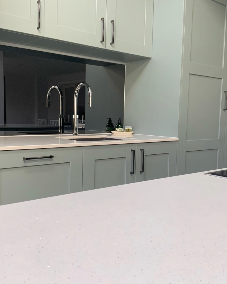 This is an example of a large classic galley open plan kitchen with shaker cabinets, green cabinets, quartz worktops, mirror splashback, integrated appliances, vinyl flooring, an island, brown floors and white worktops.