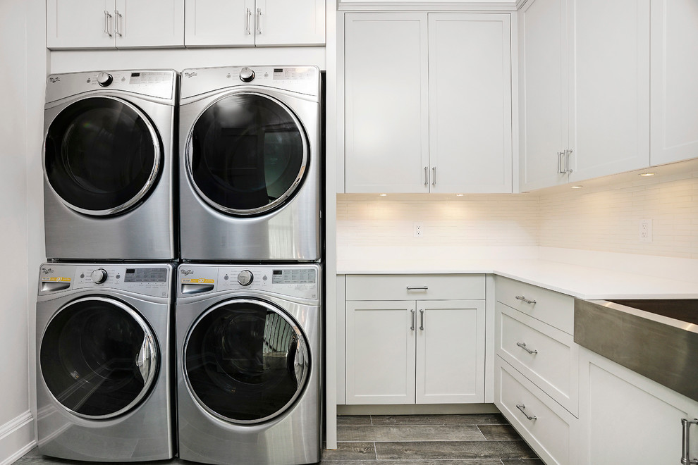 This is an example of a mid-sized transitional l-shaped dedicated laundry room in New York with a farmhouse sink, shaker cabinets, grey cabinets, quartz benchtops, white walls, porcelain floors and a stacked washer and dryer.