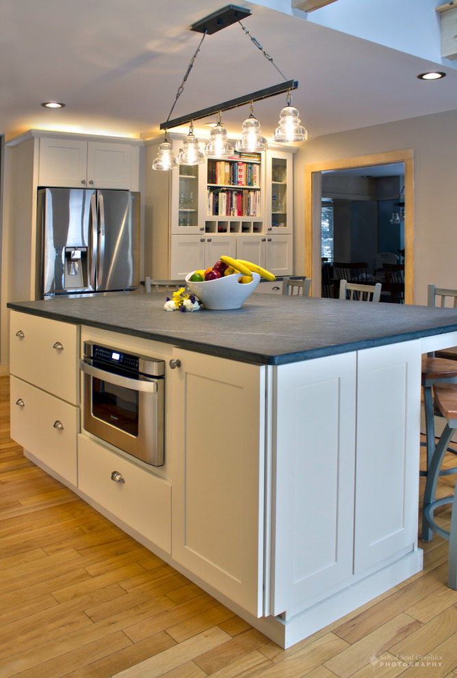 Inspiration for a large rustic l-shaped light wood floor open concept kitchen remodel in Burlington with an undermount sink, shaker cabinets, white cabinets, soapstone countertops, multicolored backsplash, glass tile backsplash, stainless steel appliances and an island