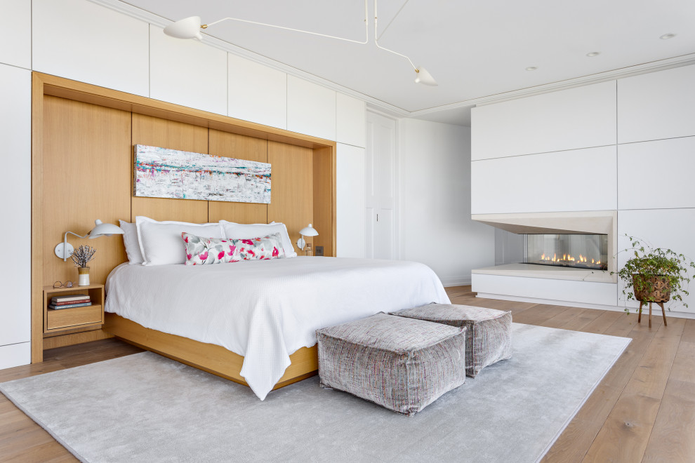This is an example of an expansive beach style master bedroom in Other with white walls, medium hardwood floors, a two-sided fireplace, a concrete fireplace surround and brown floor.
