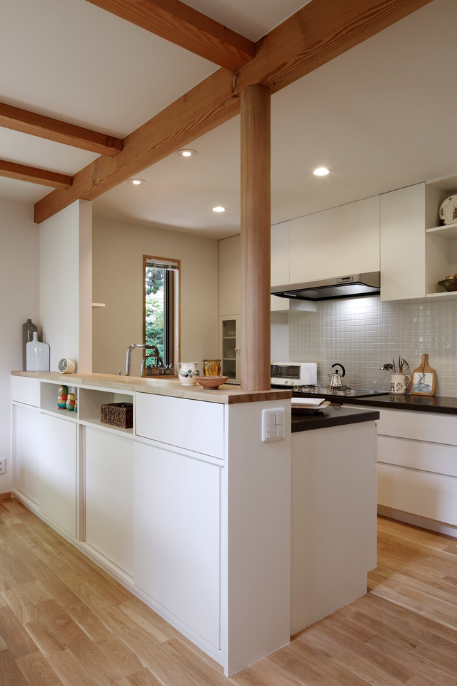 This is an example of a contemporary galley open plan kitchen in Tokyo Suburbs with flat-panel cabinets, white cabinets, white splashback, light hardwood floors, a peninsula, brown floor and brown benchtop.
