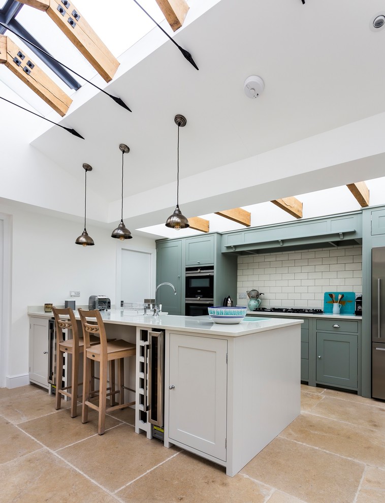 Photo of a country kitchen in Other with a farmhouse sink, recessed-panel cabinets, green cabinets, solid surface benchtops, white splashback, ceramic splashback, stainless steel appliances, limestone floors, with island, beige floor and white benchtop.
