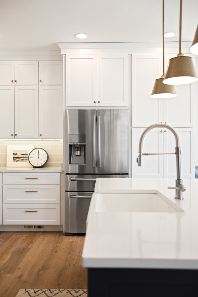 Example of a transitional l-shaped light wood floor and brown floor kitchen design with a farmhouse sink, shaker cabinets, white cabinets, quartz countertops, white backsplash, subway tile backsplash, stainless steel appliances, an island and white countertops