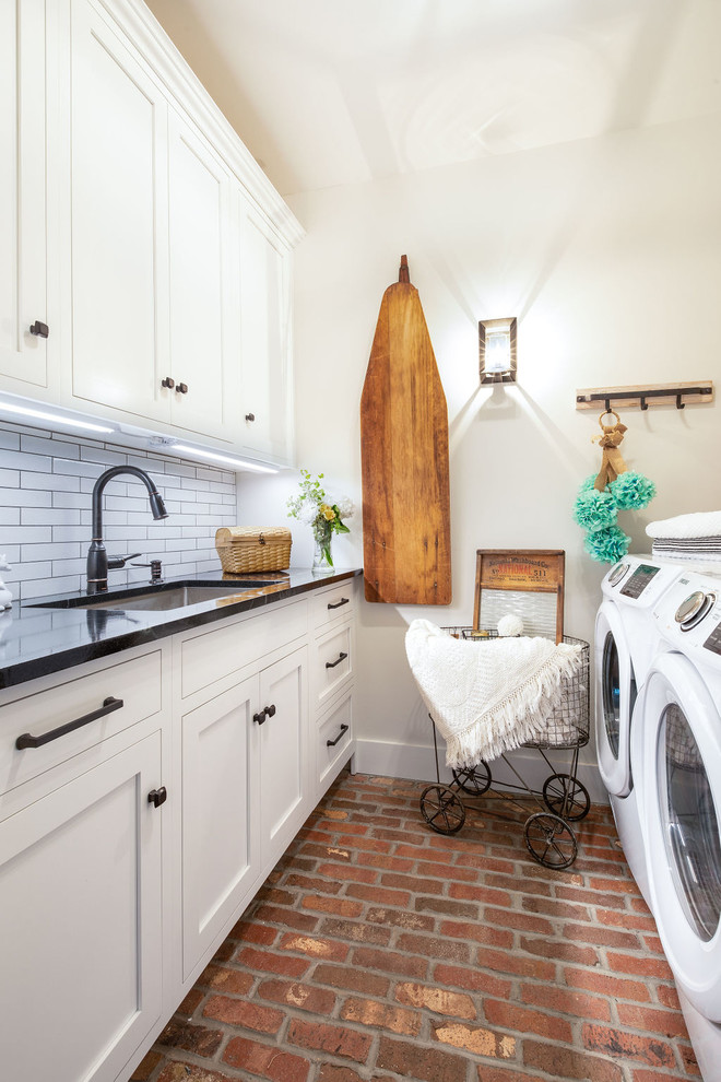 Photo of a country dedicated laundry room in Burlington with an undermount sink, shaker cabinets, white cabinets, white walls, brick floors, a side-by-side washer and dryer, red floor and black benchtop.