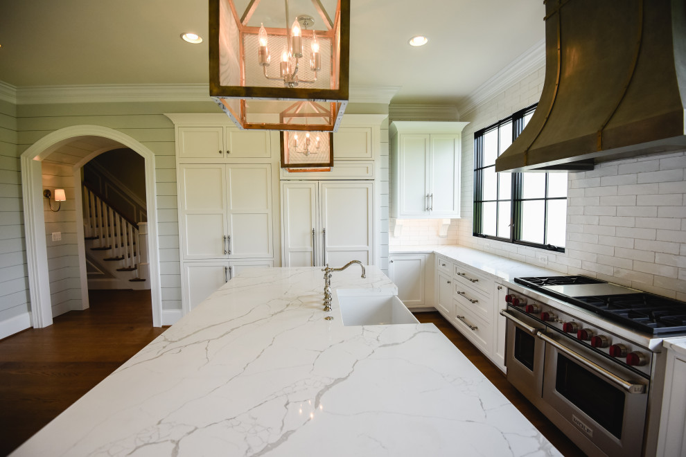 Photo of a large country u-shaped kitchen in DC Metro with a farmhouse sink, beaded inset cabinets, white cabinets, solid surface benchtops, white splashback, cement tile splashback, stainless steel appliances, medium hardwood floors, with island, brown floor and white benchtop.