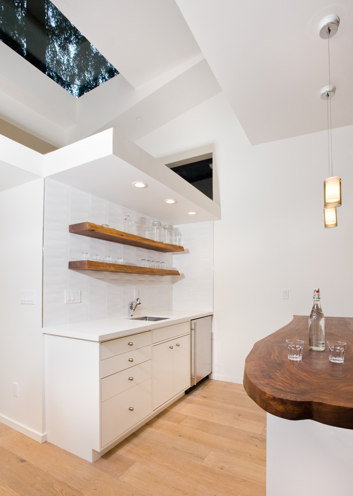 This is an example of a small modern single-wall open plan kitchen in San Francisco with open cabinets, light wood cabinets, wood benchtops, white splashback, glass tile splashback, stainless steel appliances, light hardwood floors, a peninsula and beige floor.