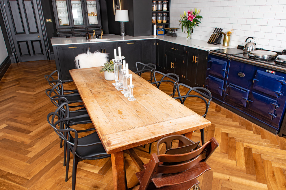 Country l-shaped eat-in kitchen in Cambridgeshire with medium hardwood floors, a farmhouse sink, recessed-panel cabinets, black cabinets, marble benchtops, white splashback, porcelain splashback, black appliances, a peninsula, brown floor and white benchtop.