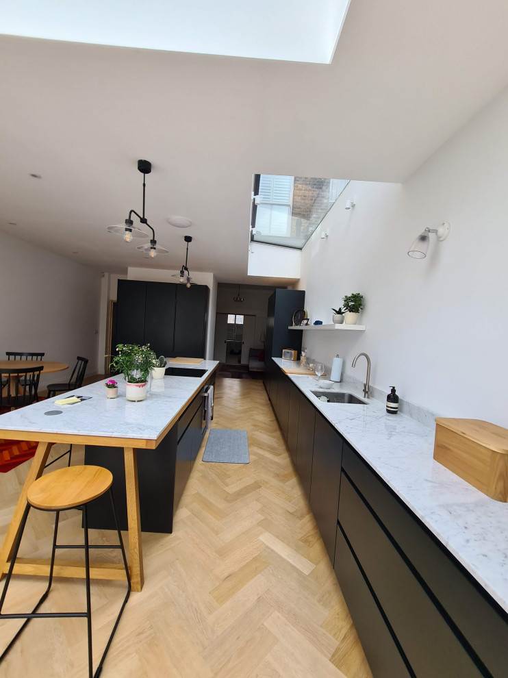 This is an example of an expansive classic grey and white l-shaped open plan kitchen in London with a built-in sink, flat-panel cabinets, black cabinets, granite worktops, white splashback, glass sheet splashback, black appliances, light hardwood flooring, an island, brown floors, white worktops, a drop ceiling and feature lighting.