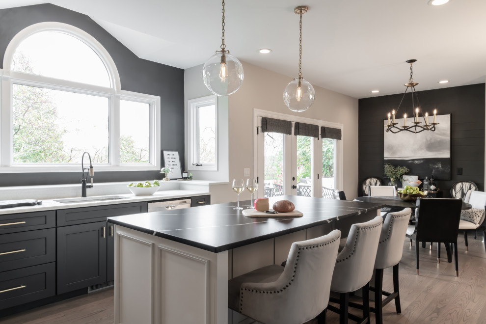 This is an example of a transitional l-shaped eat-in kitchen in St Louis with with island, an undermount sink, grey cabinets, quartz benchtops, white splashback, stainless steel appliances, medium hardwood floors, brown floor, black benchtop and shaker cabinets.