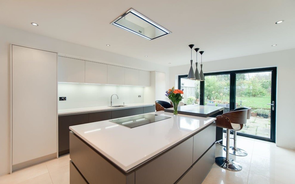 Photo of a large modern galley open plan kitchen in Kent with a drop-in sink, flat-panel cabinets, white cabinets, glass benchtops, beige splashback, glass tile splashback, stainless steel appliances, slate floors, a peninsula and beige floor.