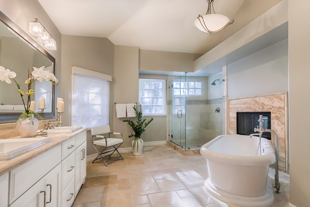 Design ideas for a large transitional master bathroom in Los Angeles with a drop-in sink, white cabinets, a freestanding tub, a corner shower, beige tile, beige walls, travertine floors, travertine, recessed-panel cabinets, marble benchtops, beige floor and a hinged shower door.