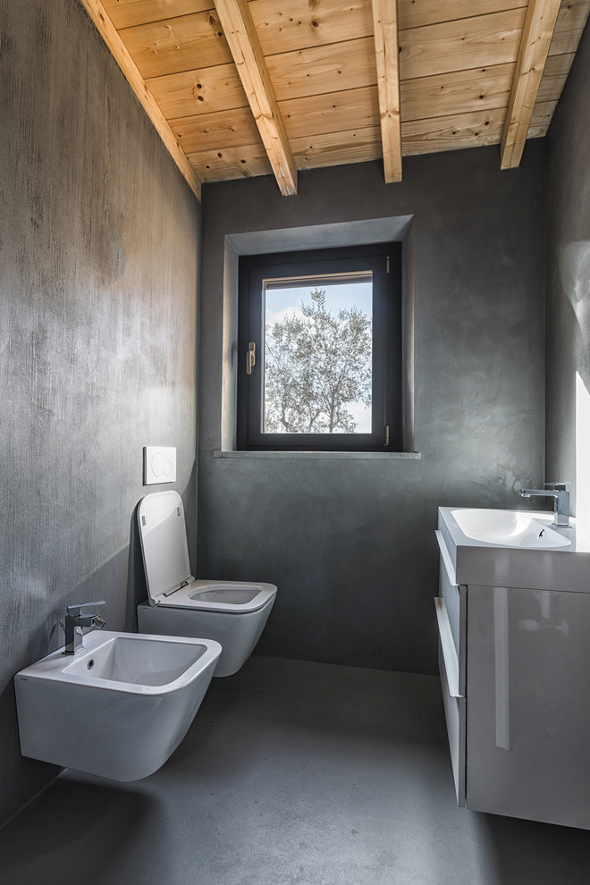 This is an example of a country powder room in Florence with flat-panel cabinets, white cabinets, a wall-mount toilet, grey walls, grey floor and concrete floors.