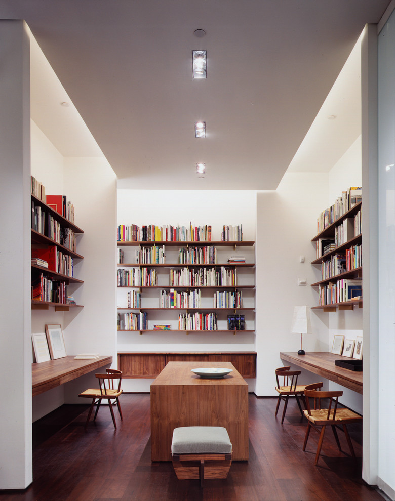 This is an example of a contemporary home office in New York with white walls, dark hardwood floors, a built-in desk and a library.