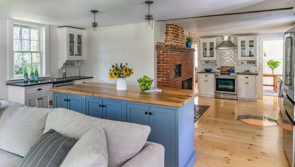 Photo of a mid-sized country l-shaped eat-in kitchen in Boston with a farmhouse sink, white splashback, light hardwood floors, beaded inset cabinets, white cabinets, granite benchtops, subway tile splashback, stainless steel appliances, with island and black benchtop.