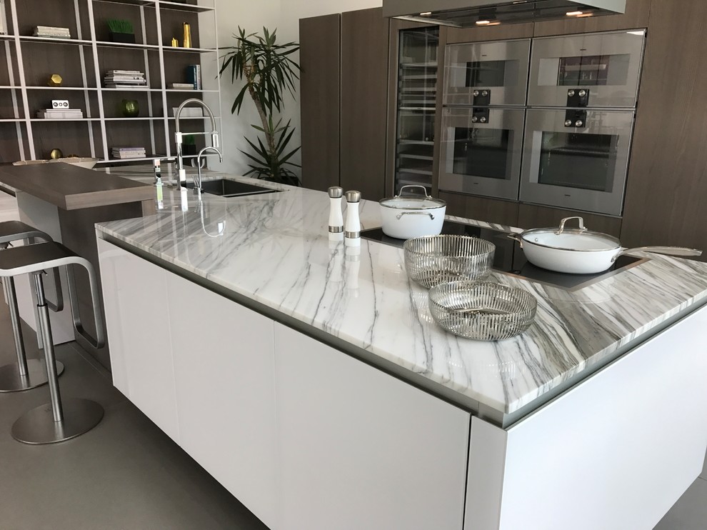 This is an example of a large contemporary u-shaped open plan kitchen in Houston with a double-bowl sink, glass-front cabinets, light wood cabinets, marble benchtops, stainless steel appliances, ceramic floors, with island, grey floor and white benchtop.