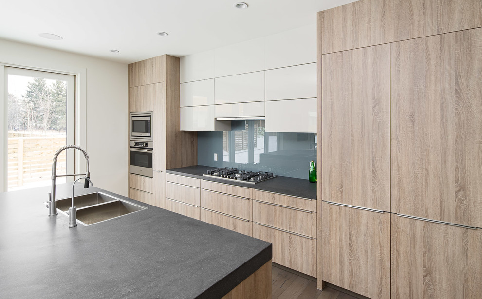 Inspiration for a large modern single-wall open plan kitchen in Calgary with a double-bowl sink, flat-panel cabinets, light wood cabinets, quartz benchtops, blue splashback, glass sheet splashback, panelled appliances, medium hardwood floors and multiple islands.