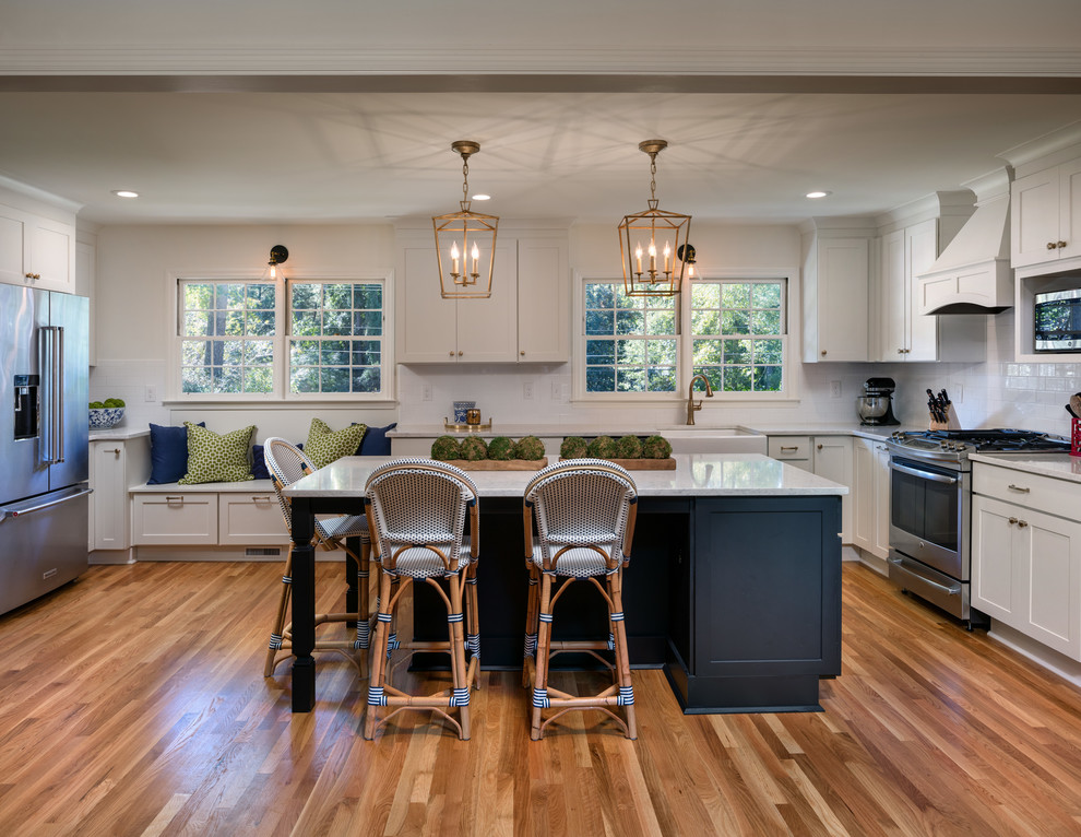 This is an example of a large transitional l-shaped eat-in kitchen in Atlanta with a farmhouse sink, recessed-panel cabinets, white cabinets, quartz benchtops, white splashback, subway tile splashback, stainless steel appliances, medium hardwood floors, with island and brown floor.
