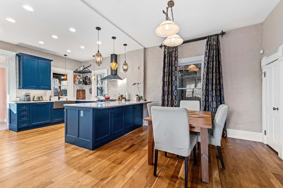 This is an example of a mid-sized eclectic u-shaped eat-in kitchen in Other with a farmhouse sink, recessed-panel cabinets, blue cabinets, quartz benchtops, grey splashback, brick splashback, coloured appliances, medium hardwood floors, a peninsula, brown floor and white benchtop.