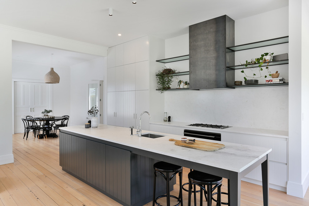 Design ideas for a mid-sized transitional galley open plan kitchen in Auckland with white cabinets, marble benchtops, white splashback, marble splashback, black appliances, light hardwood floors, with island, brown floor, white benchtop, an undermount sink and flat-panel cabinets.