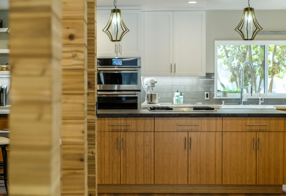 Photo of a large midcentury l-shaped eat-in kitchen in Sacramento with a farmhouse sink, flat-panel cabinets, medium wood cabinets, quartz benchtops, grey splashback, ceramic splashback, stainless steel appliances, medium hardwood floors, with island, brown floor and brown benchtop.
