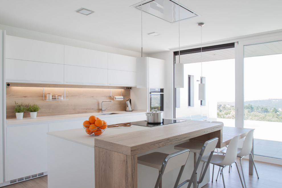 Photo of a beach style kitchen in Other with an undermount sink, flat-panel cabinets, white cabinets, beige splashback, timber splashback, black appliances, light hardwood floors, with island, beige floor and white benchtop.