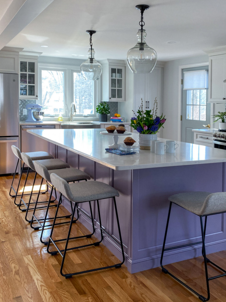 Traditional l-shaped kitchen in Boston with a belfast sink, shaker cabinets, purple cabinets, engineered stone countertops, grey splashback, ceramic splashback, stainless steel appliances, light hardwood flooring, an island and white worktops.
