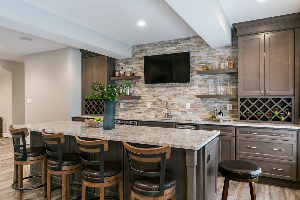 Design ideas for a large transitional single-wall seated home bar in DC Metro with brown cabinets, granite benchtops, beige splashback, stone tile splashback, laminate floors, brown floor, recessed-panel cabinets, an undermount sink and grey benchtop.