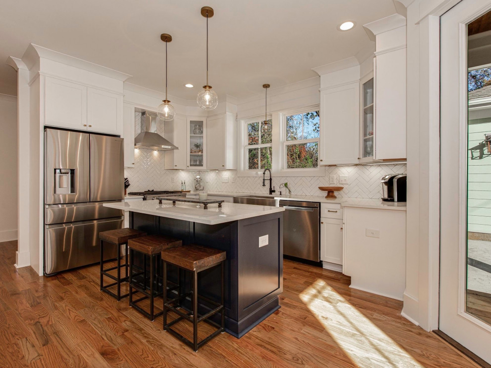 Photo of a large transitional l-shaped eat-in kitchen in Charlotte with a farmhouse sink, white cabinets, quartz benchtops, white splashback, ceramic splashback, stainless steel appliances, medium hardwood floors, with island, brown floor, white benchtop and shaker cabinets.