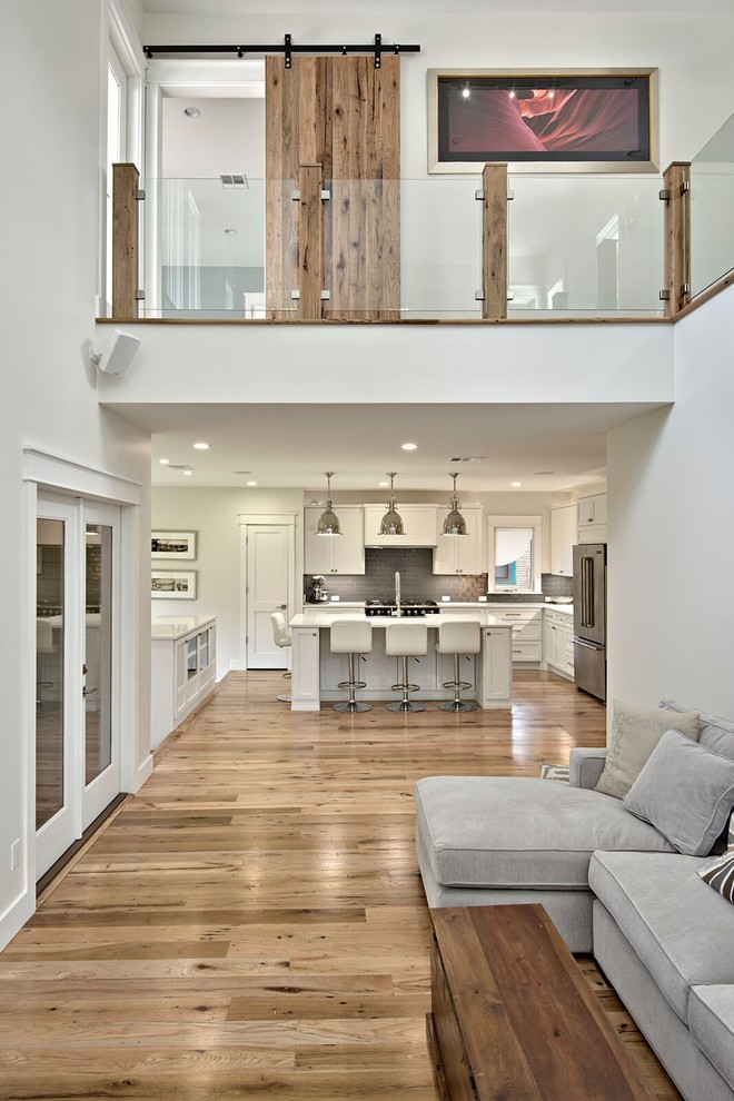 Eat-in kitchen - large transitional l-shaped light wood floor eat-in kitchen idea in Austin with a farmhouse sink, shaker cabinets, white cabinets, quartz countertops, gray backsplash, glass tile backsplash, stainless steel appliances and an island