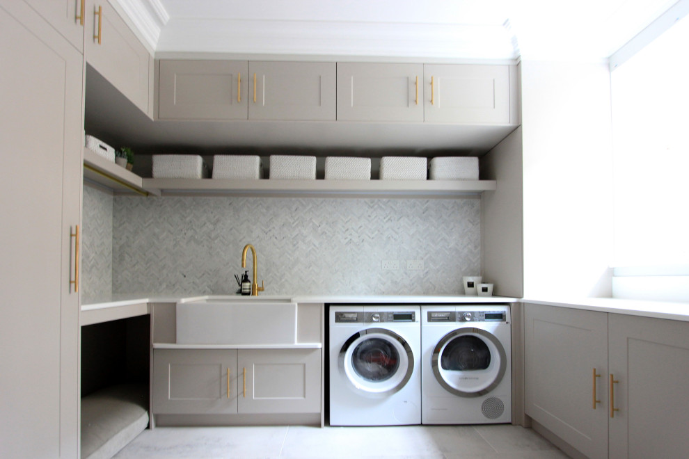 Modern inredning av en stor vita vitt liten tvättstuga, med en rustik diskho, skåp i shakerstil, grå skåp, bänkskiva i kvartsit, grått stänkskydd, stänkskydd i marmor, vita väggar, en tvättmaskin och torktumlare bredvid varandra och grått golv