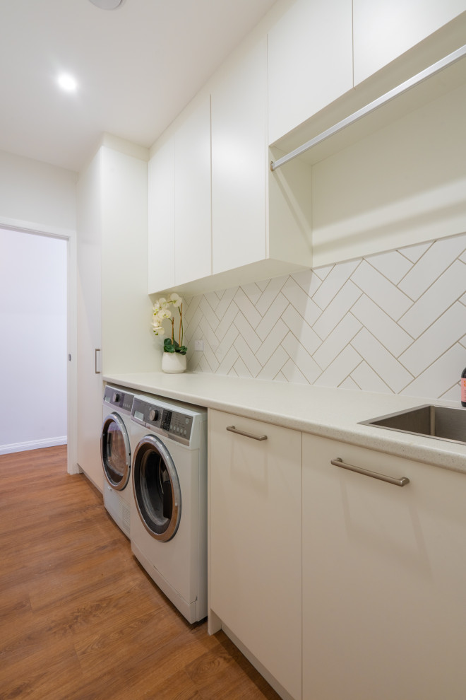 Photo of a mid-sized transitional single-wall dedicated laundry room in Auckland with a single-bowl sink, white cabinets, laminate benchtops, white splashback, subway tile splashback, white walls, laminate floors, a side-by-side washer and dryer, brown floor and white benchtop.
