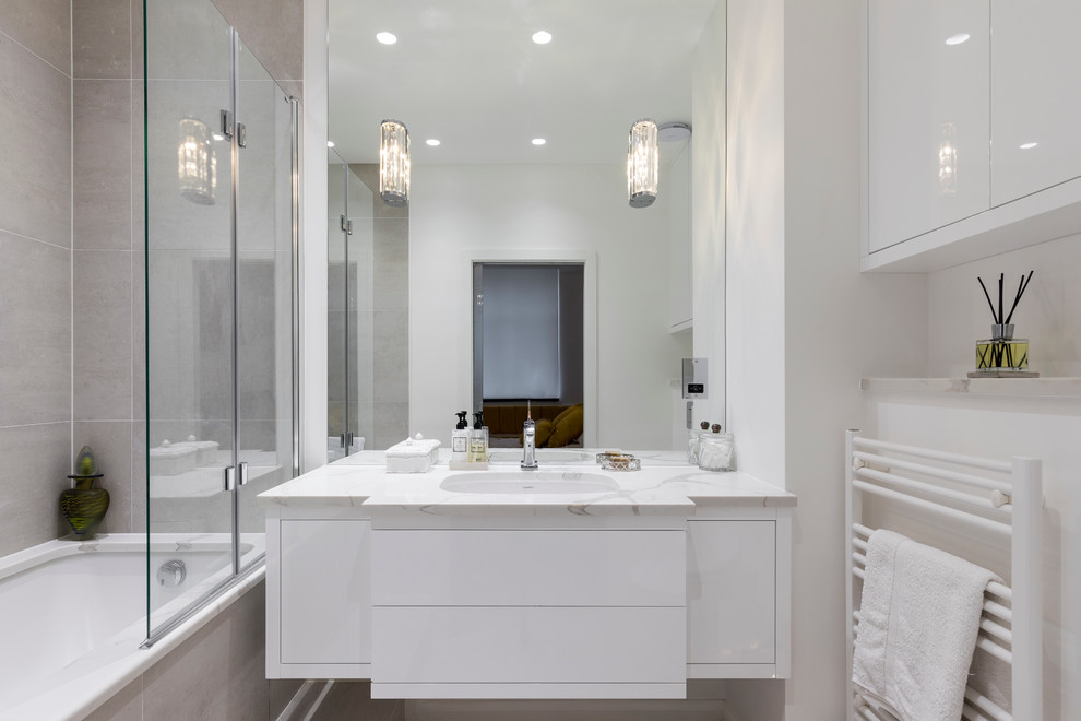 Design ideas for a mid-sized contemporary master bathroom in London with flat-panel cabinets, white cabinets, an alcove tub, a shower/bathtub combo, beige tile, gray tile, ceramic tile, white walls, ceramic floors, an undermount sink, marble benchtops, white floor, a hinged shower door and grey benchtops.