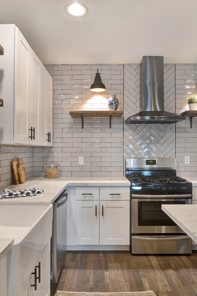 Mid-sized transitional l-shaped laminate floor and brown floor open concept kitchen photo in Houston with a farmhouse sink, shaker cabinets, white cabinets, quartz countertops, white backsplash, ceramic backsplash, stainless steel appliances, an island and white countertops