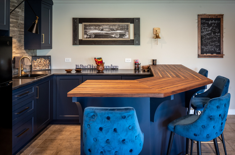 This is an example of a small transitional u-shaped wet bar in Other with a drop-in sink, shaker cabinets, blue cabinets, wood benchtops, brown splashback, stone tile splashback, ceramic floors and brown benchtop.