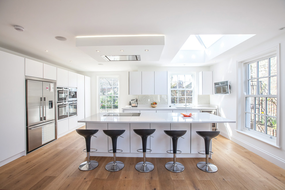 Large contemporary u-shaped kitchen in London with flat-panel cabinets, white cabinets, white splashback, stainless steel appliances, light hardwood floors and with island.