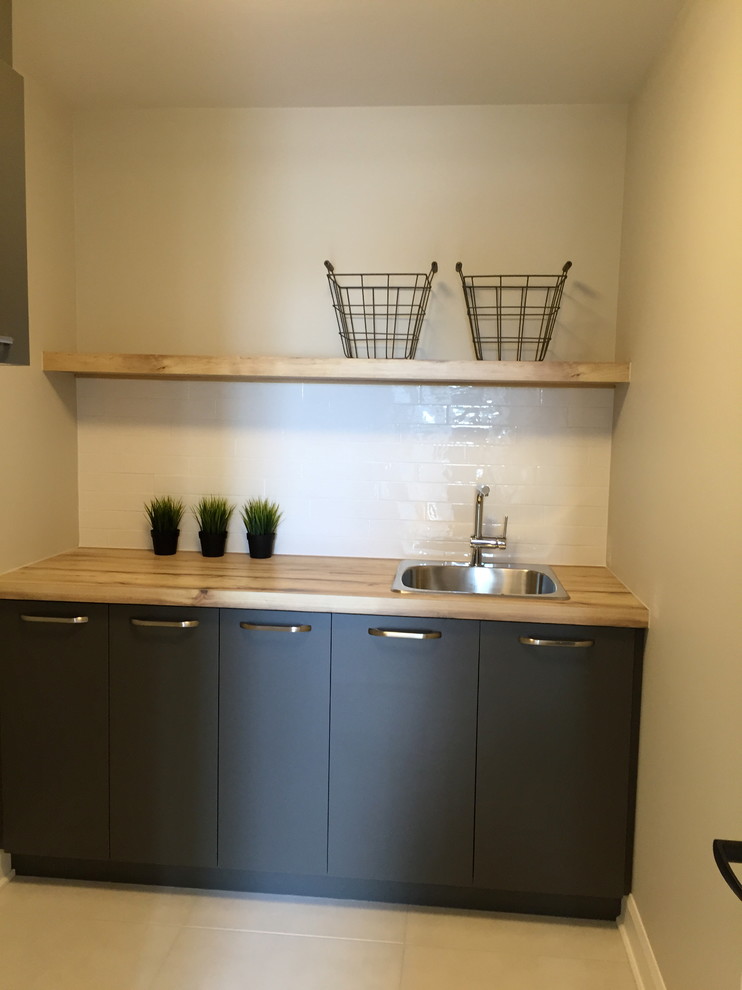 Photo of a mid-sized modern single-wall laundry room in Montreal with a drop-in sink, flat-panel cabinets, grey cabinets, beige walls, ceramic floors and beige floor.