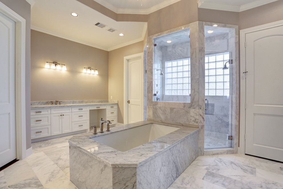 This is an example of a large transitional master bathroom in Houston with shaker cabinets, white cabinets, a freestanding tub, an alcove shower, beige tile, stone tile, beige walls, marble floors, an undermount sink, marble benchtops and grey floor.