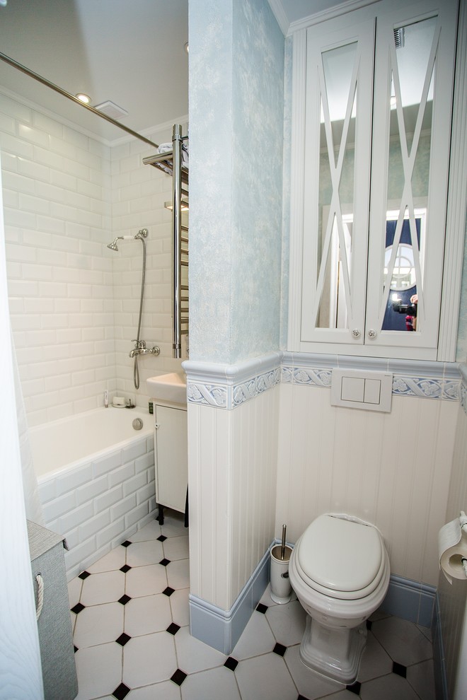This is an example of a traditional bathroom in Moscow with raised-panel cabinets, white cabinets, a drop-in tub, a shower/bathtub combo, a one-piece toilet, white tile, ceramic tile, white walls and ceramic floors.