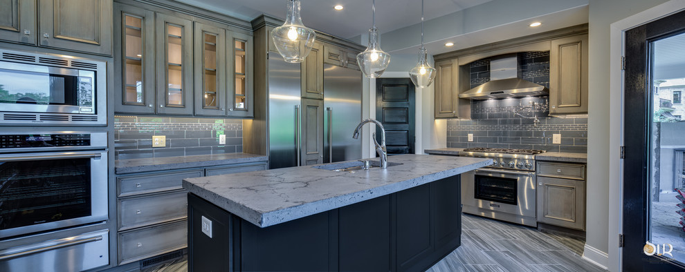Example of a transitional l-shaped medium tone wood floor and brown floor enclosed kitchen design with an undermount sink, raised-panel cabinets, distressed cabinets, soapstone countertops, gray backsplash, subway tile backsplash, stainless steel appliances and an island