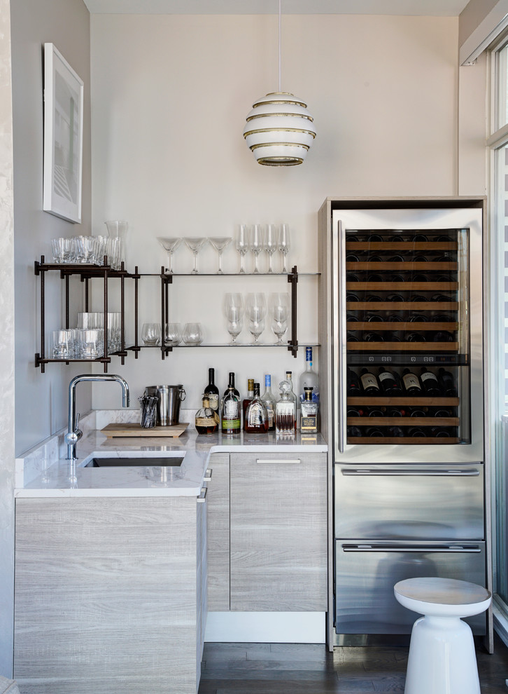 This is an example of a mid-sized contemporary l-shaped wet bar in Chicago with flat-panel cabinets, grey cabinets, white splashback, stone slab splashback, an undermount sink, dark hardwood floors, brown floor and white benchtop.