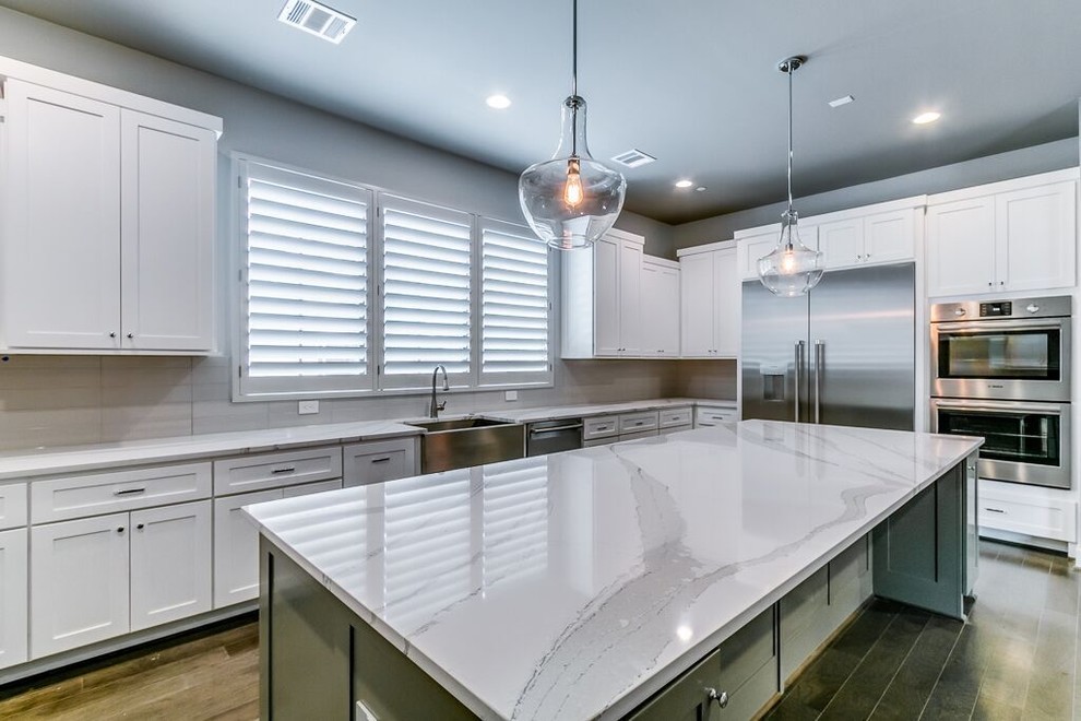 Transitional l-shaped brown floor open concept kitchen photo in Houston with a farmhouse sink, shaker cabinets, white cabinets, quartz countertops, beige backsplash, subway tile backsplash, stainless steel appliances and an island
