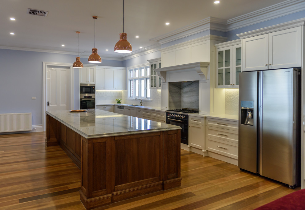 This is an example of an expansive traditional l-shaped open plan kitchen in Melbourne with a double-bowl sink, recessed-panel cabinets, dark wood cabinets, quartzite benchtops, white splashback, ceramic splashback, black appliances, medium hardwood floors and with island.