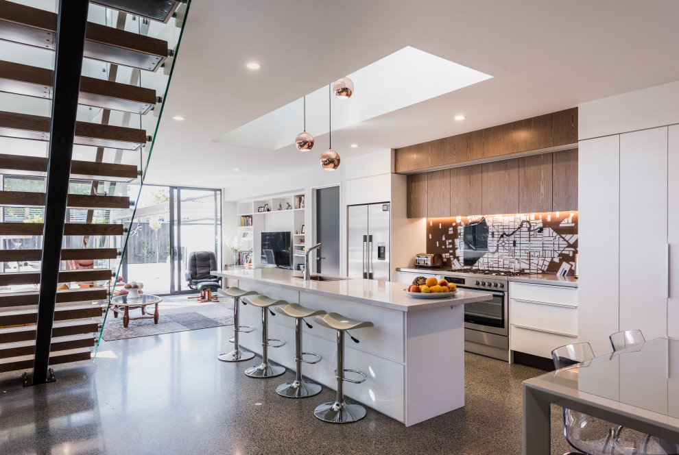 Large contemporary galley open plan kitchen in Christchurch with an undermount sink, flat-panel cabinets, white cabinets, multi-coloured splashback, stainless steel appliances, with island, grey floor and grey benchtop.