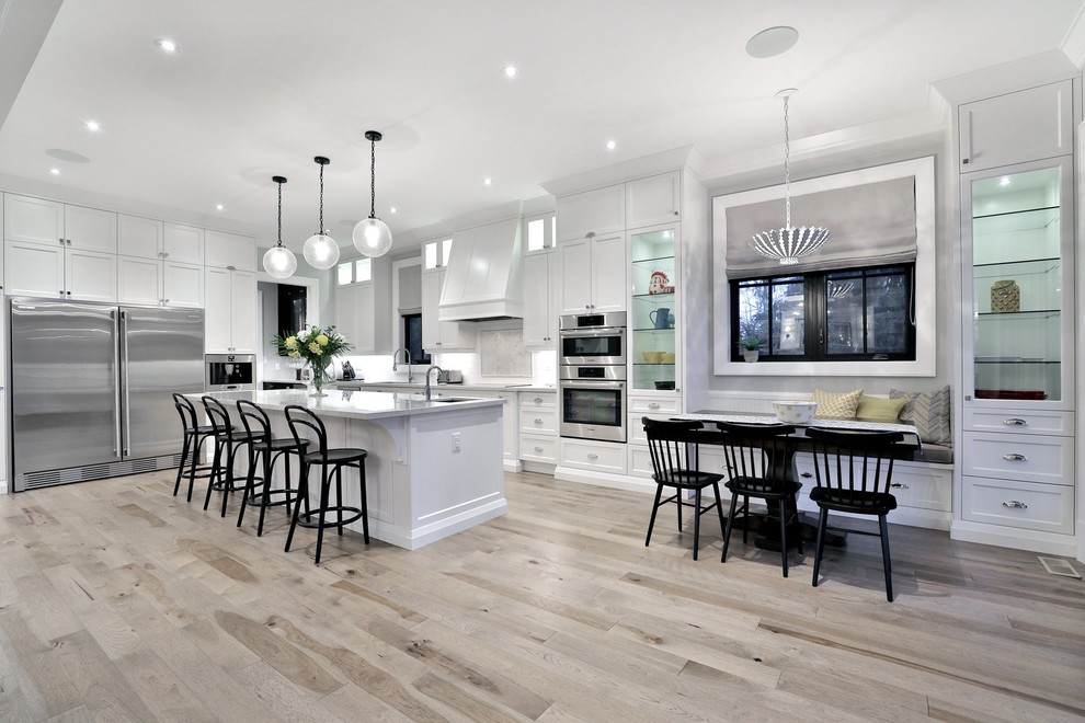 This is an example of a mid-sized transitional l-shaped eat-in kitchen with shaker cabinets, white cabinets, quartz benchtops, white splashback, glass tile splashback, stainless steel appliances, light hardwood floors and with island.