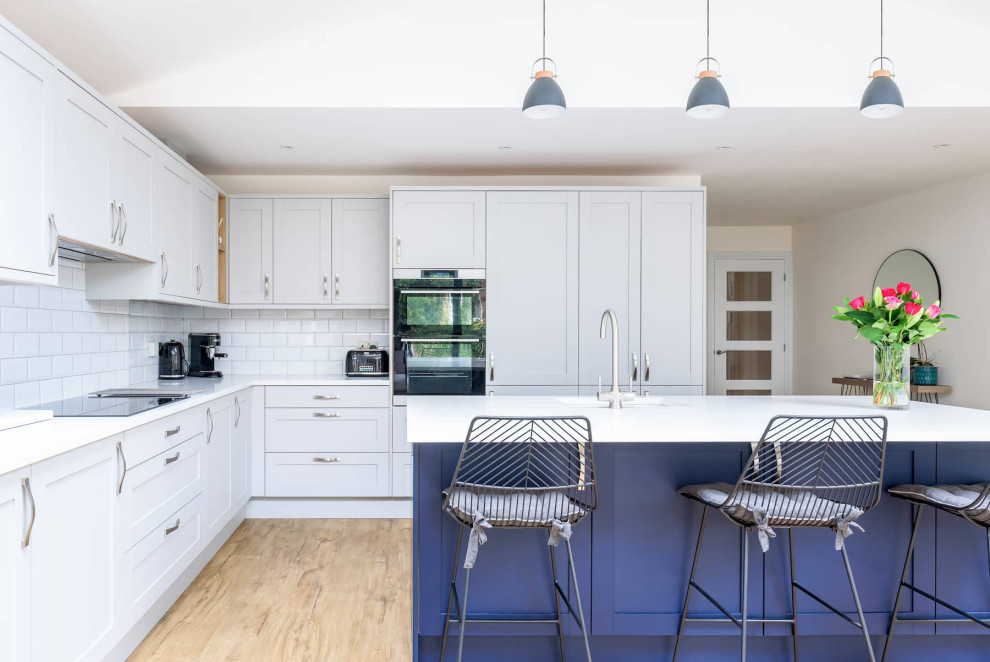 This is an example of a large traditional l-shaped open plan kitchen in Sussex with a single-bowl sink, shaker cabinets, white cabinets, quartzite benchtops, white splashback, ceramic splashback, black appliances, light hardwood floors, with island, brown floor, white benchtop and recessed.
