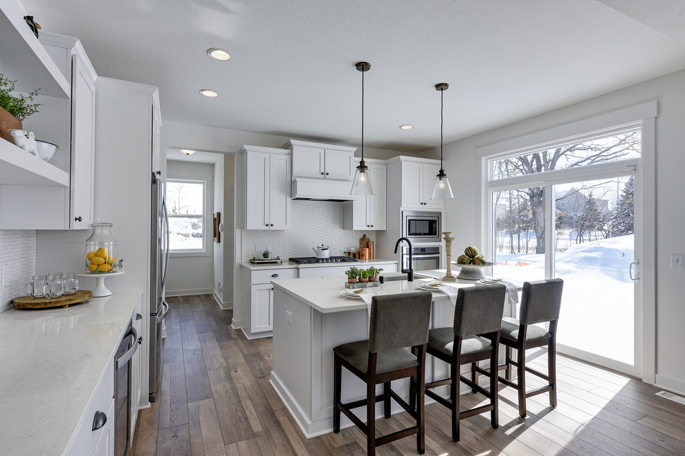 Design ideas for a small country l-shaped kitchen in Minneapolis with an undermount sink, shaker cabinets, white cabinets, quartzite benchtops, white splashback, ceramic splashback, stainless steel appliances, laminate floors, with island, white benchtop and brown floor.