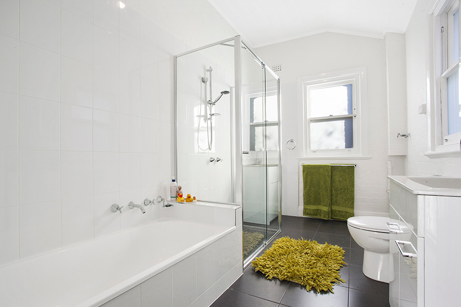 Design ideas for a mid-sized contemporary master bathroom in Melbourne with an integrated sink, furniture-like cabinets, white cabinets, solid surface benchtops, a drop-in tub, an alcove shower, a one-piece toilet, white tile, ceramic tile, white walls and ceramic floors.