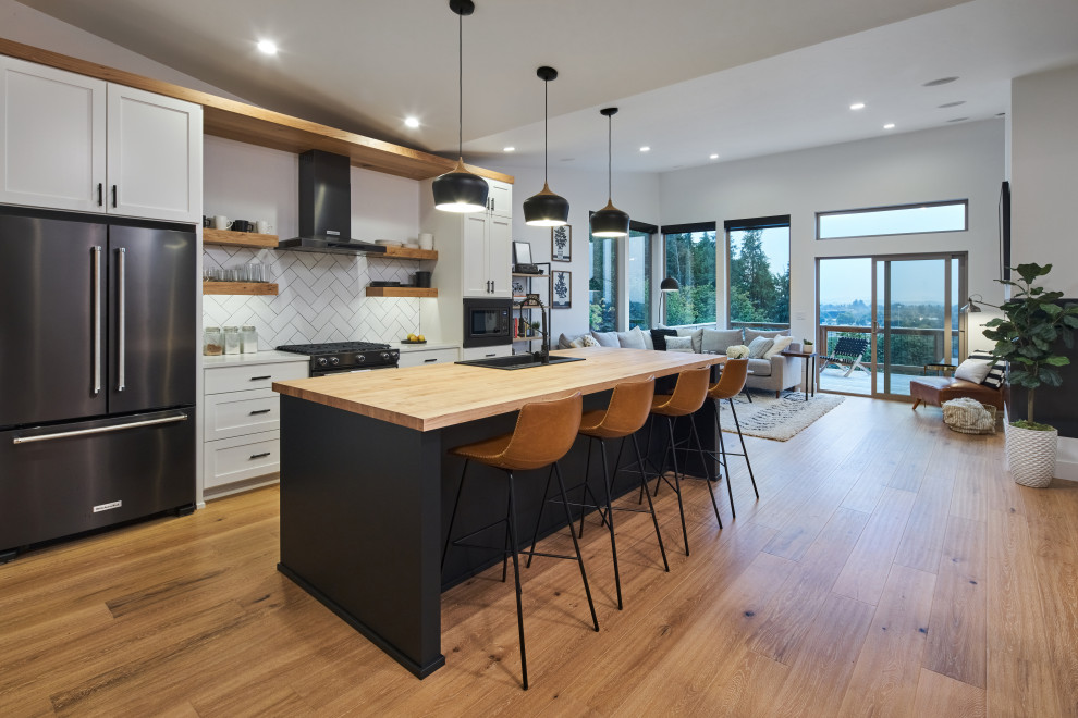 Modern inredning av ett stort brun linjärt brunt kök med öppen planlösning, med en nedsänkt diskho, skåp i shakerstil, vita skåp, träbänkskiva, vitt stänkskydd, stänkskydd i cementkakel, svarta vitvaror, mellanmörkt trägolv, en köksö och brunt golv
