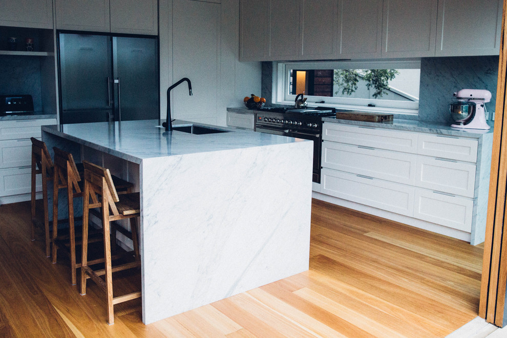 Contemporary l-shaped open plan kitchen in Sydney with white cabinets, marble benchtops, grey splashback, stone slab splashback, stainless steel appliances, medium hardwood floors, with island, an undermount sink and recessed-panel cabinets.