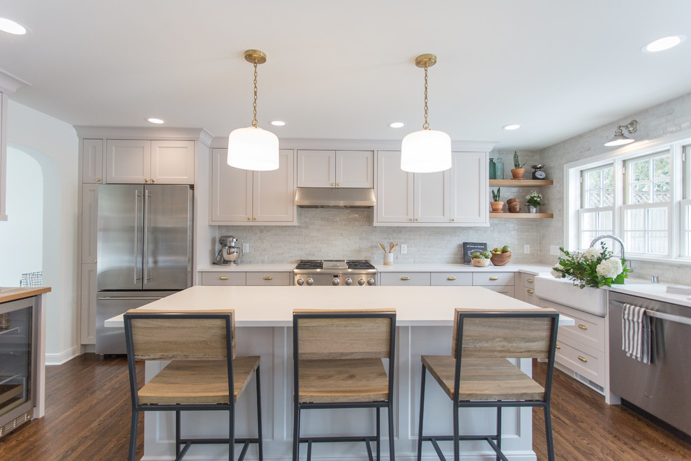 This is an example of a traditional eat-in kitchen in Minneapolis with a farmhouse sink, recessed-panel cabinets, grey cabinets, quartz benchtops, white splashback, terra-cotta splashback, stainless steel appliances, dark hardwood floors, with island, brown floor and white benchtop.